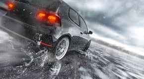 Voiture en mouvement sur route mouillée, de jour, lumineuse ; gros plan possible sur un pneu qui projette de l'eau. Texte alternatif : « Voiture roulant sur une route mouillée sous la pluie »